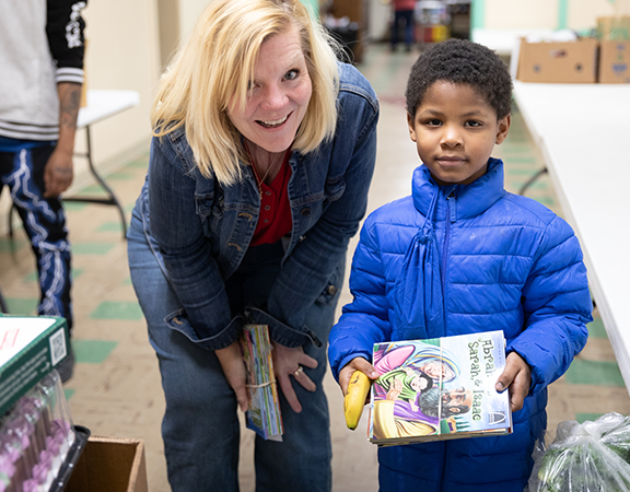 A smiling blonde woman in a denim jacket leans down next to a young boy in a bright blue puffer jacket who is holding a children's book titled "Abraham, Sarah, & Isaac" and a banana in a community distribution room. A smiling blonde woman in a denim jacket leans down next to a young boy in a bright blue puffer jacket who is holding a children's book titled "Abraham, Sarah, & Isaac" and a banana in a community distribution room.