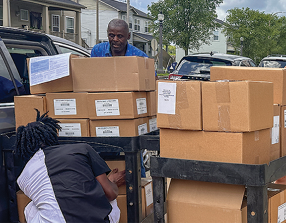 An older Black man in a blue shirt unloads heavy cardboard boxes from a van onto a rolling cart, while another person crouches in the foreground helping to stack the boxes outdoors in a residential area. An older Black man in a blue shirt unloads heavy cardboard boxes from a van onto a rolling cart, while another person crouches in the foreground helping to stack the boxes outdoors in a residential area.